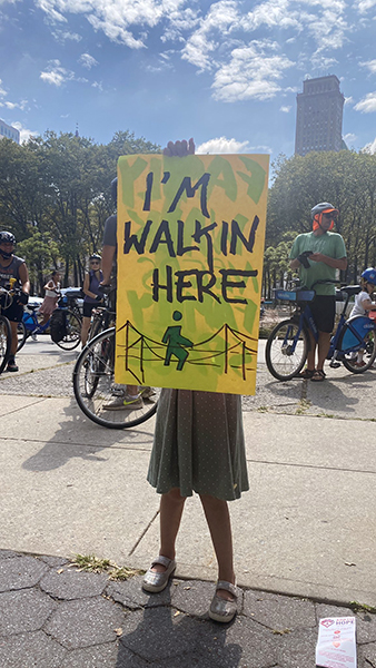 Kid holds up a colorful sign that says I'm walkin here with a drawing of a pedestrian crossing a bridge